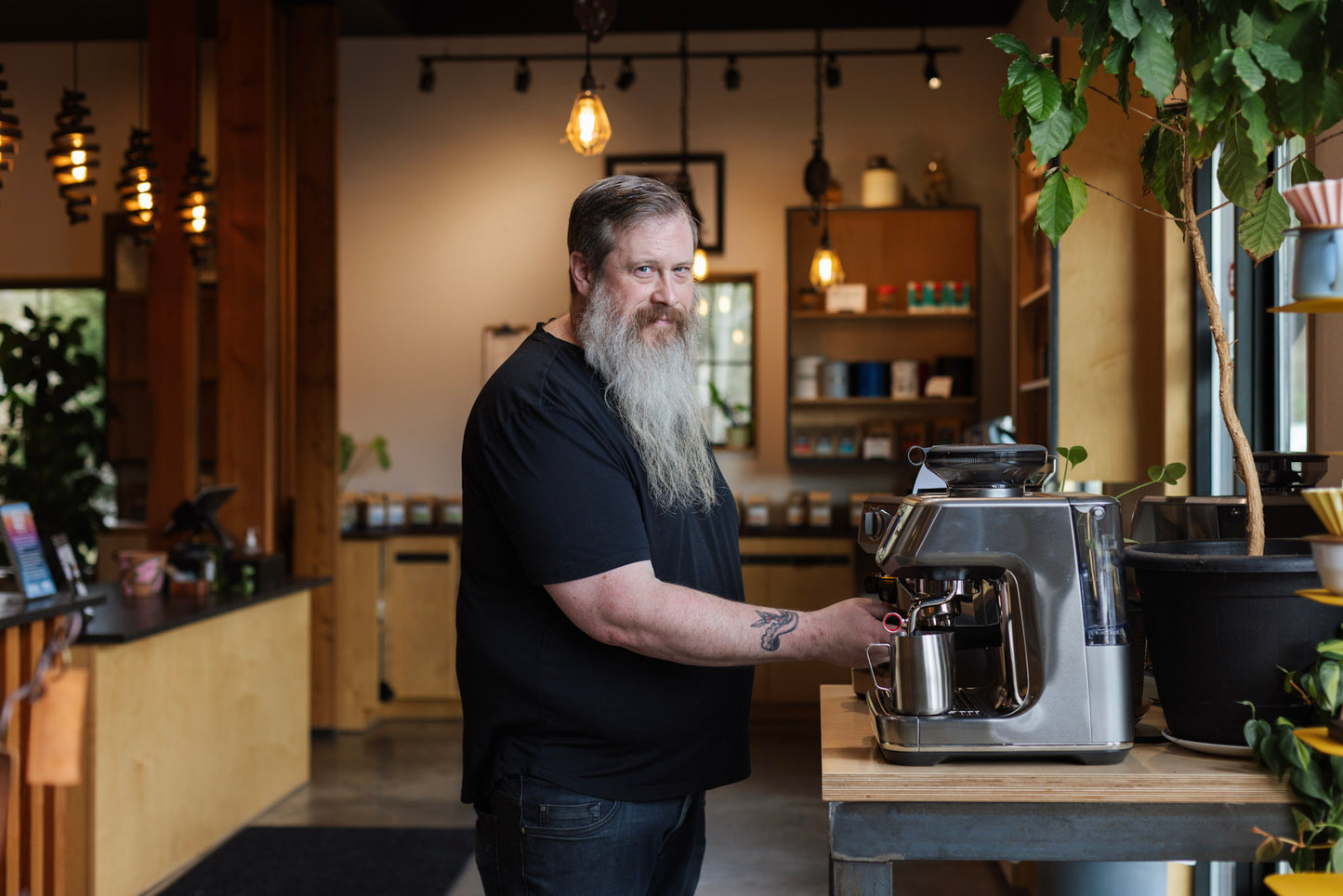 Man with a beard standing in a coffee shop next to a coffee machine.