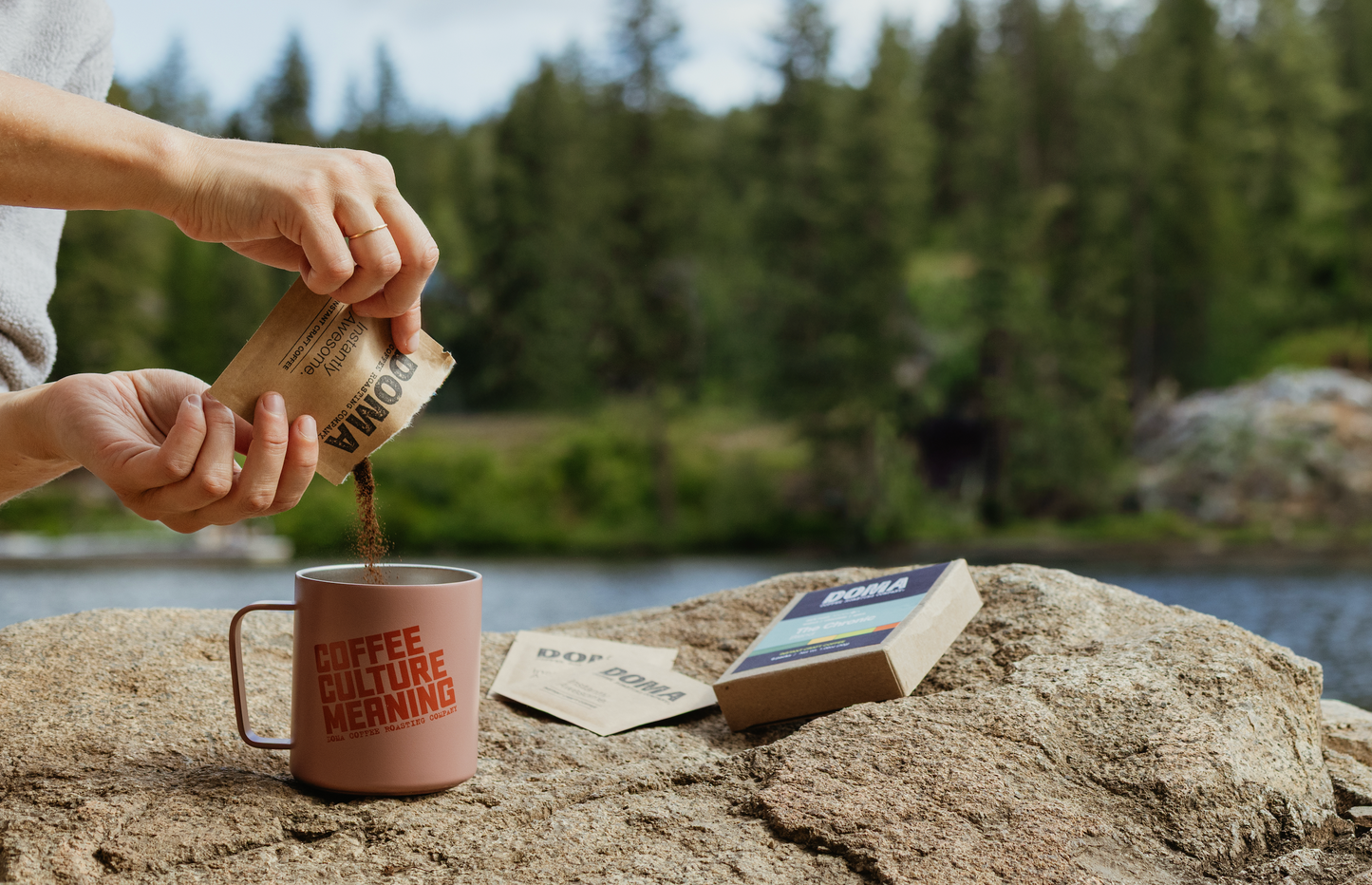 Person pouring coffee into a mug with a scenic background of trees and water.
